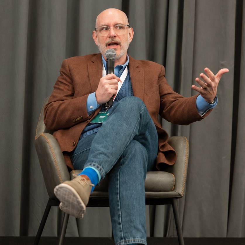 David speaking on a panel, holding a microphone and gesturing.